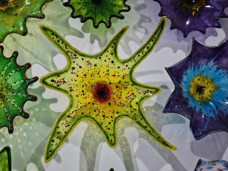 May include: A collection of colorful, star-shaped glass art pieces. The central piece is yellow with red and green accents, surrounded by green and purple pieces. The glass art has a textured appearance and is mounted on a white surface.