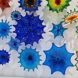 May include: A collection of colorful, spiky glass art pieces mounted on a white wall. The pieces vary in size and color, including shades of blue, orange, yellow, and green. The art has a unique, textured appearance, resembling sea urchins or starbursts.