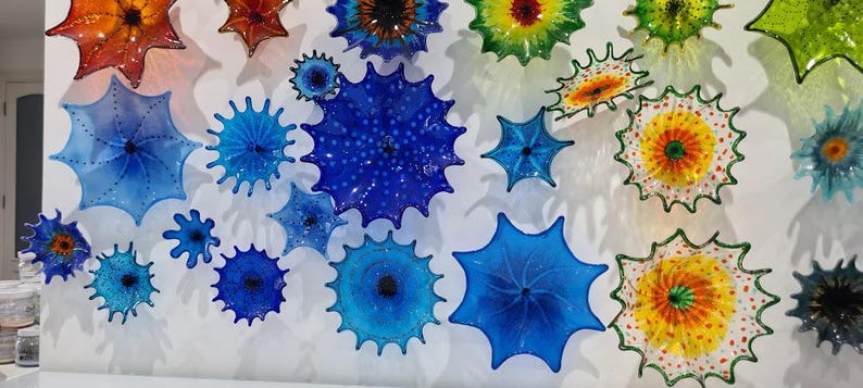 May include: A collection of colorful, spiky glass art pieces mounted on a white wall. The pieces vary in size and color, including shades of blue, orange, yellow, and green. The art has a unique, textured appearance, resembling sea urchins or starbursts.