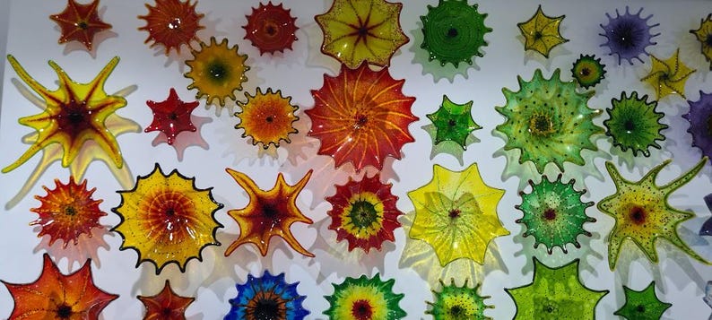 May include: A collection of colorful, starburst-shaped glass art pieces in various sizes and hues, including red, orange, yellow, green, and blue. The pieces are mounted on a white wall, creating a vibrant display.