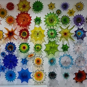 May include: A vibrant wall art installation featuring numerous colorful, starburst-shaped glass pieces. The artwork showcases a spectrum of colors, including red, orange, yellow, green, blue, and purple, creating a visually striking display. The pieces vary in size and design.