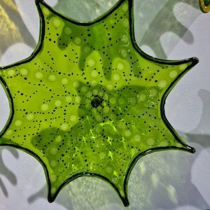 May include: A lime green, star-shaped glass art piece with black trim and a pattern of small black dots. The glass is translucent, allowing light to pass through. Other colorful glass pieces are visible in the background.