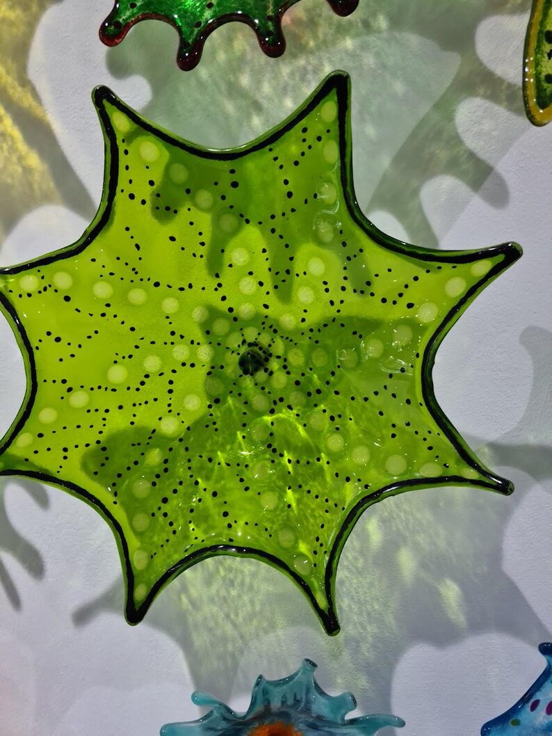 May include: A lime green, star-shaped glass art piece with black trim and a pattern of small black dots. The glass is translucent, allowing light to pass through. Other colorful glass pieces are visible in the background.