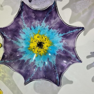 May include: A decorative glass art piece in the shape of a flower. The flower has a deep purple outer layer, transitioning to a light blue and yellow center. Black speckles add detail. The piece is mounted on a white wall.