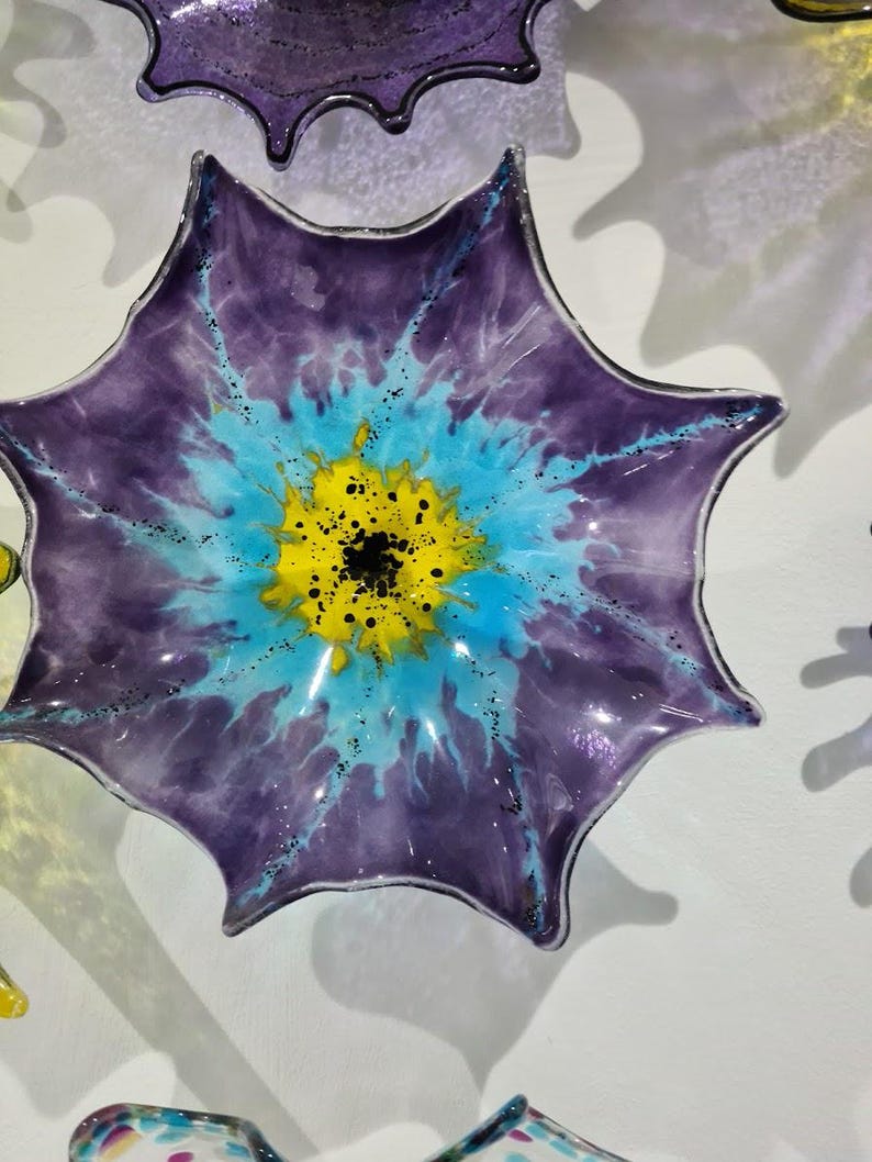 May include: A decorative glass art piece in the shape of a flower. The flower has a deep purple outer layer, transitioning to a light blue and yellow center. Black speckles add detail. The piece is mounted on a white wall.