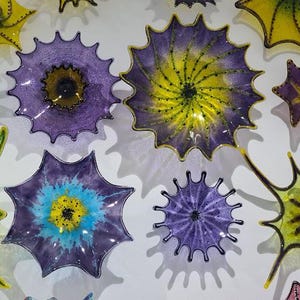 May include: A collection of colorful, spiky glass art pieces in shades of yellow, green, purple, and blue. The pieces are mounted on a white wall, creating a vibrant and artistic display. The shapes vary, resembling stars and floral designs.