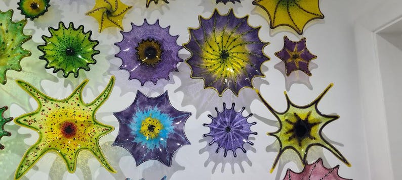 May include: A collection of colorful, spiky glass art pieces in shades of yellow, green, purple, and blue. The pieces are mounted on a white wall, creating a vibrant and artistic display. The shapes vary, resembling stars and floral designs.