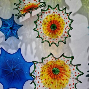 May include: A collection of colorful glass art pieces. The pieces are in the shape of flowers or starbursts, with vibrant colors including yellow, orange, green, and blue. Some have a dotted pattern.
