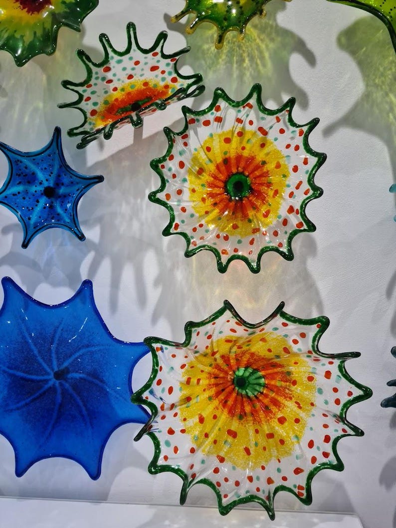 May include: A collection of colorful glass art pieces. The pieces are in the shape of flowers or starbursts, with vibrant colors including yellow, orange, green, and blue. Some have a dotted pattern.