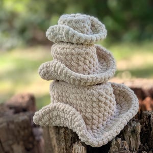 Pode incluir: Uma pilha de três chapéus de cowboy de crochê bege. Os chapéus são feitos de um fio macio e texturizado e são empilhados uns sobre os outros. Os chapéus estão em um toco de árvore desgastado, com um fundo verde desfocado.