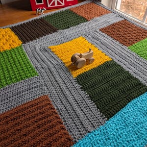 Könnte beinhalten: Eine gehäkelte Babydecke mit Farm-Motiv. Die Decke besteht aus verschiedenen farbigen Quadraten, darunter grau, braun, gelb, grün und blau. Ein kleiner Holzspielzeugtraktor liegt auf der Decke.