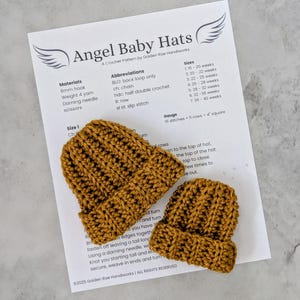 May include: Two small, mustard-coloured crochet baby hats sit on a printed pattern sheet. The sheet reads "Angel Baby Hats" and includes instructions, abbreviations, and sizes for the hats. The hats are handmade and have a ribbed texture.