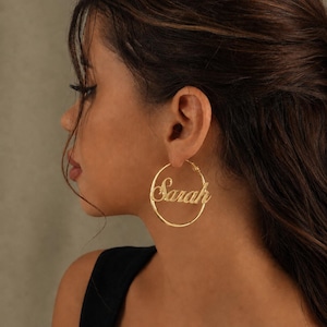 Op de afbeelding: Een goudkleurige oorring met de naam Sarah in een cursief lettertype geïntegreerd in het midden van de cirkelvormige ring. De oorbel is vervaardigd uit gepolijst metaal met een reflecterend oppervlak en een dun, cirkelvormig draadontwerp.