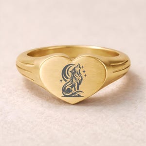 May include: A gold-colored signet ring with a heart-shaped face. The face features a detailed gray illustration of a howling wolf under a crescent moon and stars. The ring has a smooth, polished finish and a thick band with subtle ribbed detailing.