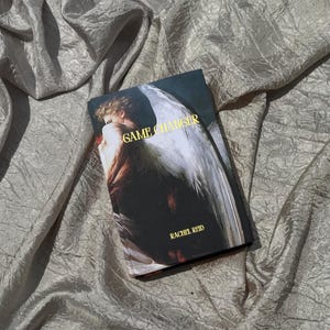 May include: A book titled "GAME CHANGER" by Rachel Reid, featuring an angel illustration on the cover. The book is set against a backdrop of a textured, silvery fabric.