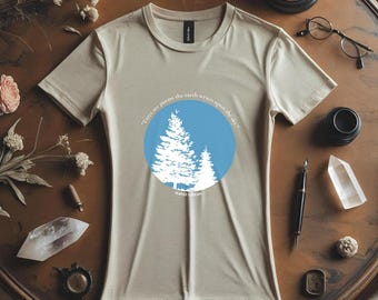 May include: Beige t-shirt with a round neck featuring a blue circle with white tree silhouettes and the quote "Trees are poems the earth writes upon the sky." by Kahlil Gibran. The shirt is displayed on a wooden table with decorative items.