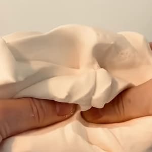 Peut inclure: Gros plan sur une matière douce, blanc cassé, tenue et manipulée par deux mains. La matière semble souple et lisse, avec des plis et des plis visibles.