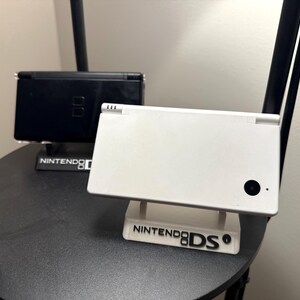 May include: A white Nintendo DS handheld game console on a stand with the text "Nintendo DS". A black Nintendo DS is visible in the background. The consoles are on a dark gray surface.