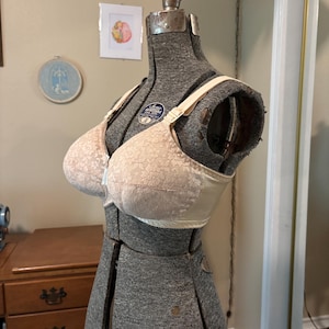 May include: A vintage beige bra with a delicate lace overlay, displayed on a gray dress form. The bra features adjustable straps and a front closure. The overall design is classic and elegant, showcasing the intricate details of the lace.