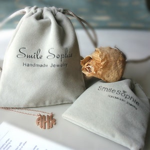 May include: Two gray velvet jewelry pouches with the text "Smile Sophie Handmade Jewelry" printed on them. A gold necklace with a monogram pendant is lying on top of one of the pouches.