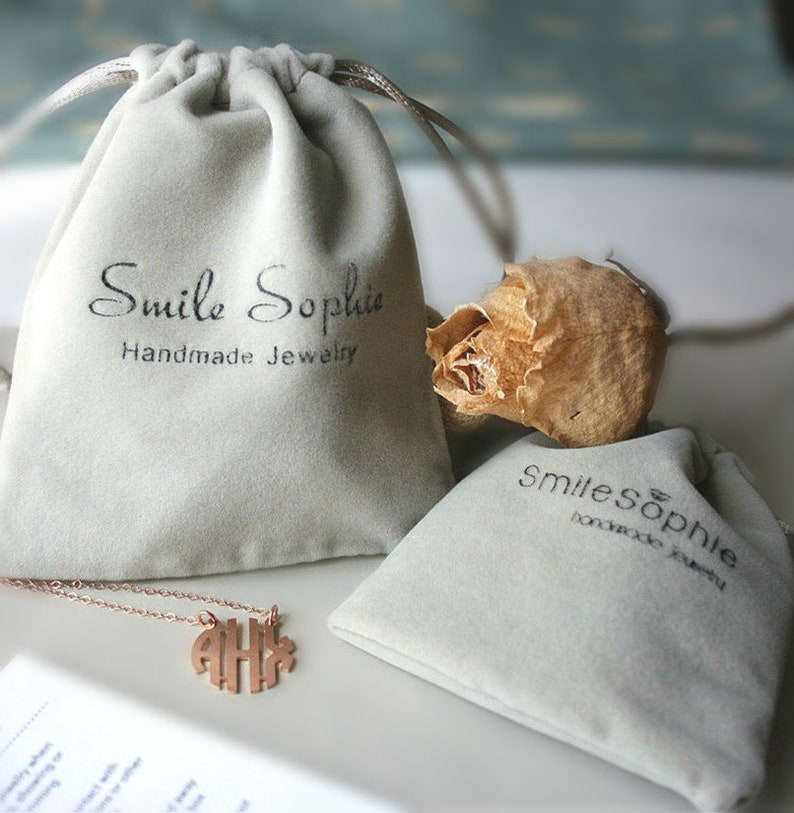 May include: Two gray velvet jewelry pouches with the text "Smile Sophie Handmade Jewelry" printed on them. A gold necklace with a monogram pendant is lying on top of one of the pouches.