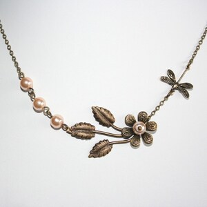 May include: A delicate bronze necklace with a floral design featuring a small pearl in the center of the flower. The necklace also has a dragonfly charm and a row of small pink pearls.