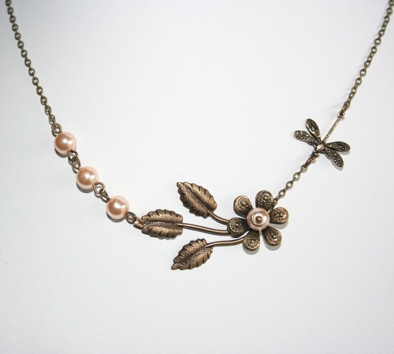 May include: A delicate bronze necklace with a floral design featuring a small pearl in the center of the flower. The necklace also has a dragonfly charm and a row of small pink pearls.