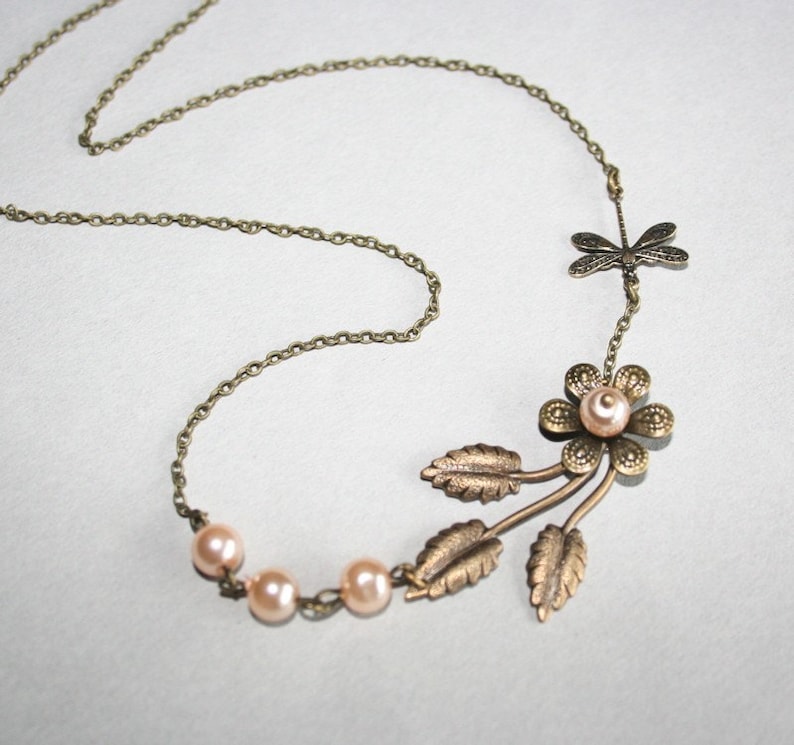 May include: A delicate gold necklace with a floral pendant featuring a pearl center and three leaves. The necklace also includes a dragonfly charm and three small pearls.