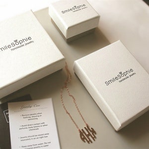 May include: Three white boxes with the text "SmileSophie handmade jewelry" on them. A gold necklace with a monogram pendant is in the center of the image. A small card with the text "Jewelry Care" and instructions for caring for jewelry is in the lower left corner of the image.