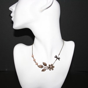 May include: A delicate gold necklace with a dragonfly charm and a floral design featuring a small flower and leaves. The necklace is adorned with small, pale pink pearls.