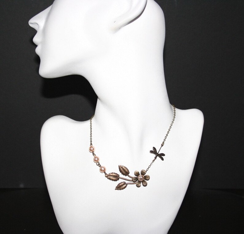 May include: A delicate gold necklace with a dragonfly charm and a floral design featuring a small flower and leaves. The necklace is adorned with small, pale pink pearls.
