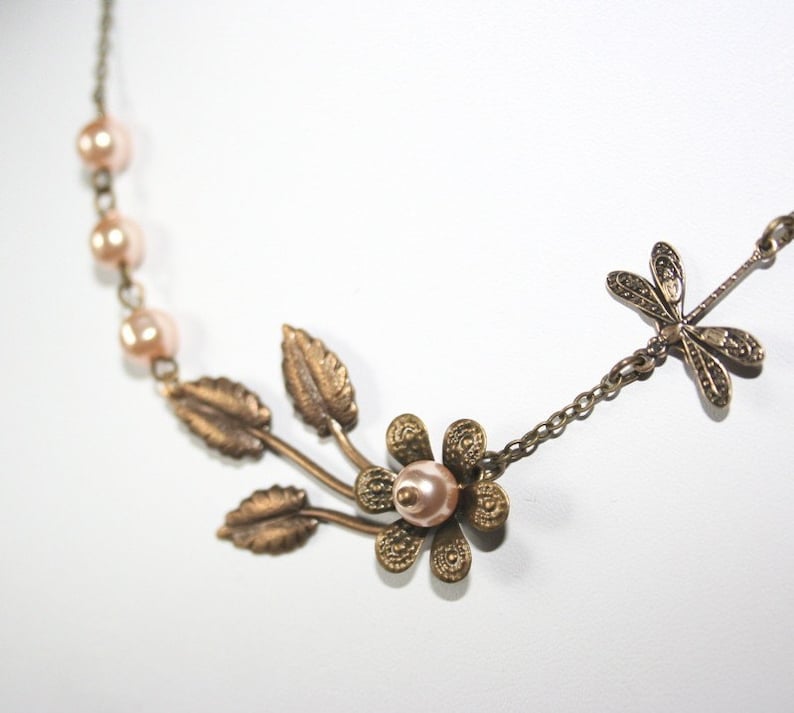 May include: A delicate necklace with a bronze dragonfly charm, a flower with a pearl center, and leaves. The necklace is made with a chain and small pearl beads.