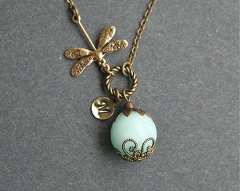 May include: A delicate gold necklace with a dragonfly charm and a light blue glass bead with an ornate metal frame. The bead has a letter 'M' charm attached to it.