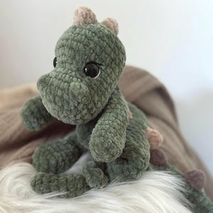 Könnte beinhalten: Ein handgefertigtes, grünes gehäkeltes Dinosaurier-Plüschtier mit großen schwarzen Augen und kleinen braunen Hörnern. Das Stofftier hat ein strukturiertes Aussehen und sitzt auf einer weißen, flauschigen Oberfläche.