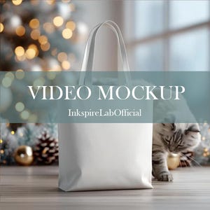 May include: A white tote bag with handles, set against a blurred background of Christmas decorations and a cat. The text "VIDEO MOCKUP" and "InkspireLabOfficial" are displayed on the image.
