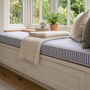 Puede incluir: Un asiento de ventana con un cojín a rayas azules y blancas, una manta de punto color crema y cojines decorativos. Una pequeña planta en maceta y una taza están en el alféizar. El asiento de ventana es parte de un mueble blanco.