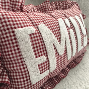 May include: A red and white gingham pillow with the name "EMILY" embroidered in white, featuring a ruffled edge. The pillow is a decorative item, likely for a bedroom or living space.