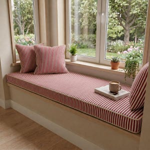 May include: A window seat featuring a red and white striped cushion and two matching pillows. A book and a mug of coffee rest on the cushion. The window offers a view of a garden with plants.