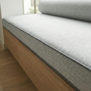 May include: A light gray upholstered bench with a wooden base. The bench features a rectangular seat with a contrasting gray trim and a rolled bolster pillow. The wooden base has a natural wood grain finish.
