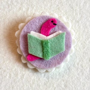 May include: A felt bookworm pin. The pin shows a pink worm reading a green book on a lilac background. The lilac circle has a white scalloped edge. The bookworm pin is a charming accessory.