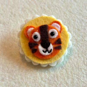 May include: A handmade felt tiger face pin. The tiger has orange and brown stripes, black eyes, and a black nose. The tiger is centered on a yellow felt circle, which is surrounded by a white scalloped edge.