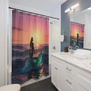 Puede incluir: Cortina de ducha con la silueta de una sirena en las olas al atardecer. La sirena tiene una cola turquesa, y el cielo pasa del rosa al naranja. La cortina está en un baño con gabinetes blancos y una encimera de mármol.