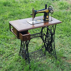 Op de afbeelding: Antiek zwarte Singer naaimachine met een houten tafelblad en een lade. De machine staat op een zwart metalen onderstel met ingewikkelde details.