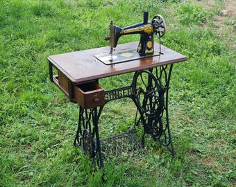 May include: Antique black Singer sewing machine with a wooden table top and a drawer. The machine is on a black metal stand with intricate details.