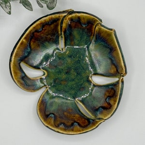 May include: A handcrafted ceramic dish with a unique, organic shape. The dish features a blend of green, brown, and blue-green glaze, creating a marbled effect. The edges have a darker, more defined color. The dish has cut-out sections.