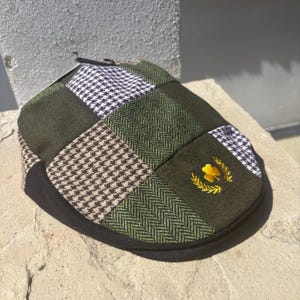 May include: A flat cap featuring a patchwork design of olive green solid panels, brown and white houndstooth, and green herringbone patterns. The cap includes a small embroidered yellow shamrock emblem surrounded by a golden laurel wreath.