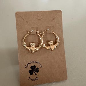 Könnte beinhalten: Goldfarbene Creolen-Ohrringe mit Claddagh-Design, bestehend aus Herz, Krone und Händen. Die Ohrringe sind auf einer braunen Karte mit dem Text "Handmade by Niamh" und einem Kleeblatt abgebildet.