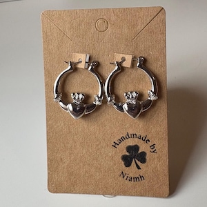 May include: Silver Claddagh hoop earrings, with a heart, crown, and hands design. The earrings are presented on a brown card with the text "Handmade by Niamh" and a shamrock.