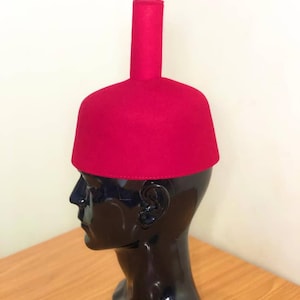 May include: A bright red fez hat, featuring a cylindrical crown and a rounded, flat top. The hat is displayed on a black mannequin head. The hat's bold colour and shape make it a distinctive accessory.