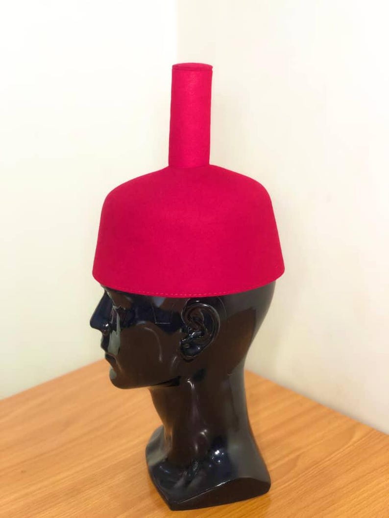 May include: A bright red fez hat, featuring a cylindrical crown and a rounded, flat top. The hat is displayed on a black mannequin head. The hat's bold colour and unique shape make it stand out.